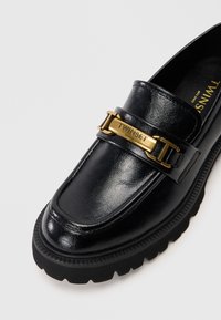 Loafers din piele neagră cu o talpă masivă neagră, prevăzuți cu un accent din lanț auriu și o curea cu marcaj, textură netedă și design clasic.
