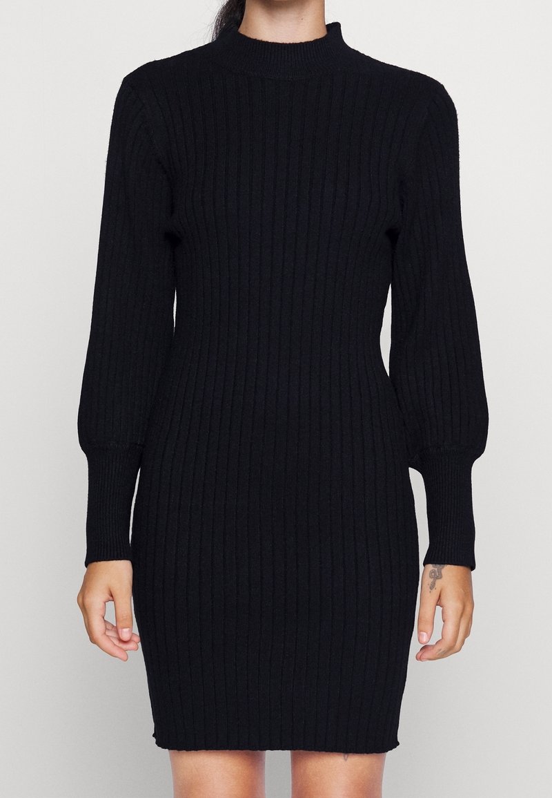 Femme portant une robe noire en maille côtelée ajustée avec de longues manches bouffantes et un col montant, debout devant un fond uni.