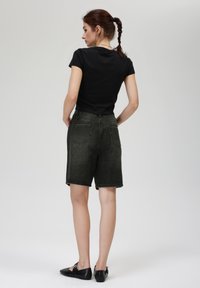Chemise noire à manches courtes, shorts en denim vert foncé avec une coupe légèrement ample, comportant deux poches arrière et un ourlet brut. Chaussures noires.