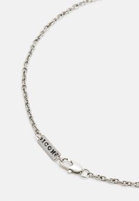 Colar de corrente prateada com elos pequenos, apresentando uma etiqueta retangular texturizada marcada com "ICON" e um fecho em metal. Acabamento liso e polido.