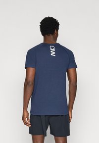 Navy zilā sporta t-krekl, kas izgatavots no mīksta auduma, ar apaļu kakla izgriezumu un vertikālu baltu logo uz aizmugures. Īsas piedurknes, pieguļošs dizains.