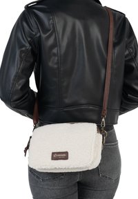 Une personne portant une veste en cuir noire et un jean gris porte un sac en fourrure blanche en bandoulière avec une sangle brune et un écusson logo visible.