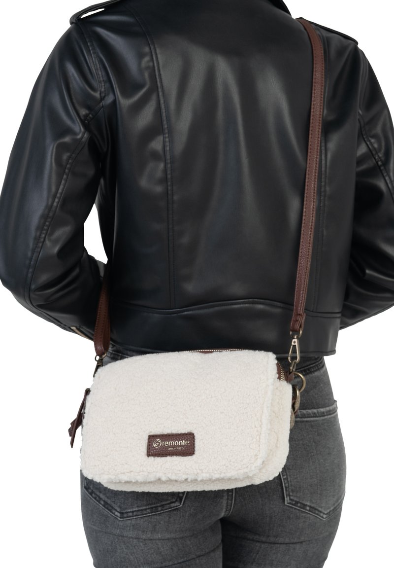 Une personne portant une veste en cuir noire et un jean gris porte un sac en fourrure blanche en bandoulière avec une sangle brune et un écusson logo visible.
