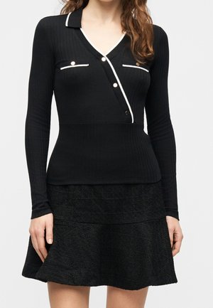 Femme portant un haut noir à manches longues avec une bordure blanche et des boutons dorés, associé à une jupe noire texturée évasée.