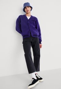 Cárdigan púrpura con botones negros y rayas, llevado sobre una camisa blanca. Combinado con jeans oscuros y zapatillas negras con suelas blancas.