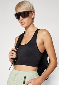 Μαύρο αθλητικό crop top με ριγωτή υφή, συνδυασμένο με ανοιχτό πράσινο παντελόνι με κορδόνι. Το μοντέλο φοράει μεγάλα μαύρα γυαλιά ηλίου και κρατάει μια μαύρη τσάντα πλάτης.