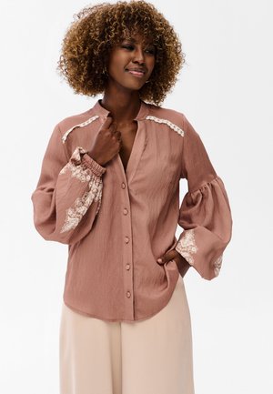 Kobieta z kręconymi włosami w zakurzonym różowym, zapinanym na guziki bluzce z koronkowymi detalami na ramionach i bufkami z kwiatowym haftem na rękawach.