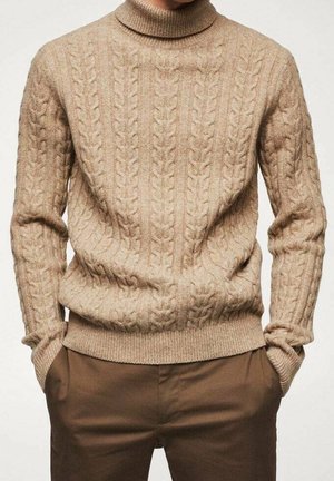 Pull à col roulé beige en maille torsadée, porté avec un pantalon marron, mains dans les poches, montré des épaules aux hanches.
