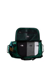Sac de voyage vert avec un compartiment zippé contenant trois chemises pliées (noire, grise, blanche) et une bouteille d'eau bleue sur le côté.
