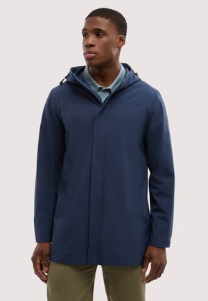 Uomo che indossa una giacca con cappuccio blu navy sopra una polo azzurra e pantaloni verde oliva, che guarda alla sua sinistra contro uno sfondo semplice.