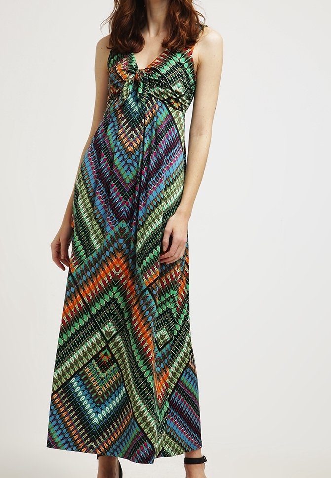 Femme portant une robe longue sans manches à motif géométrique coloré, avec un col en V et un détail froncé à l'avant, debout devant un fond uni.