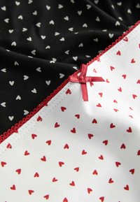 Gros plan sur un tissu noir et blanc à petits motifs de cœurs, bordé d'un liseré rouge avec un petit nœud rouge à la couture.