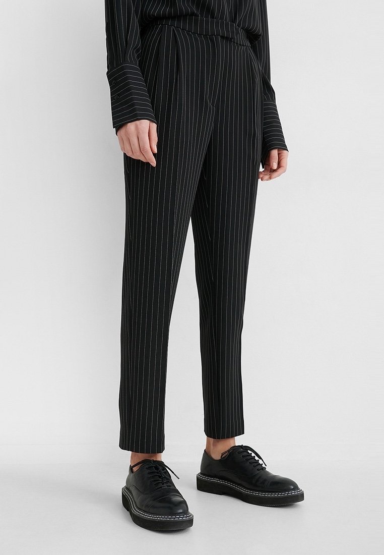 Personne portant un pantalon tailleur noir à fines rayures et un haut à manches longues assorti, ainsi que des chaussures en cuir noires à lacets, debout sur un sol blanc.