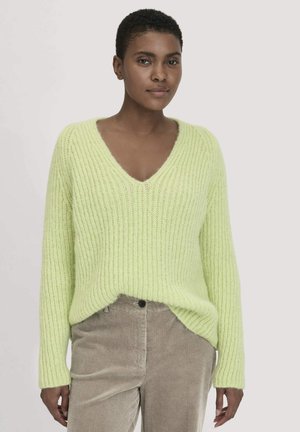 V-Ausschnitt, übergroßer Pullover in hellgrün mit gerippter Textur. Kombiniert mit beigen Cordhosen, die einen Knopf und subtile Rillen aufweisen.
