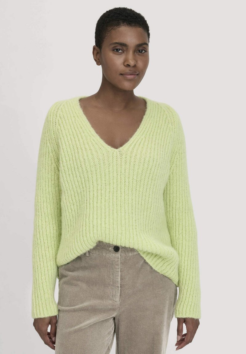 V-Ausschnitt, übergroßer Pullover in hellgrün mit gerippter Textur. Kombiniert mit beigen Cordhosen, die einen Knopf und subtile Rillen aufweisen.