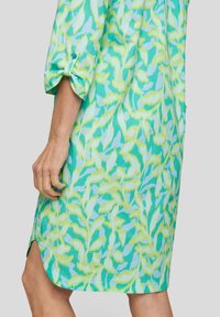 Kurzärmliges Kleid mit einem Blattmuster in Türkis- und Limettengrüntönen. Weicher Stoff mit leicht abgerundetem Saum und seitlichen Taschen.