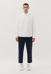 Camicetta bianca a maniche lunghe con chiusura a bottoni, abbinata a pantaloni blu navy e sneakers bianche, su uno sfondo chiaro e semplice.