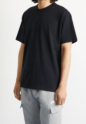 T-shirt noir à manches courtes en tissu coton ; présente un petit logo Nike noir sur la poitrine ; porté avec un pantalon de survêtement gris.