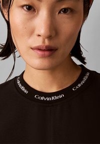 Κοντινό πλάνο ενός ατόμου με κοντά σκούρα μαλλιά που φοράει μαύρη μπλούζα με λογότυπο Calvin Klein στο γιακά.