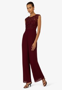 Burgunderfarbener Spitzeneinteiler mit tailliertem Oberteil und weitem Bein. Kurze Ärmel und filigrane Blumenverzierungen im oberen Bereich. Elegante schwarze Absatzschuhe.