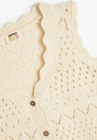 Krämfärgad stickad väst med scallopkrage, knappstängning och öppet mönster med diamant- och chevronformer.