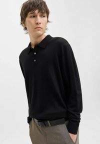 Jaunas vyras su ruda šukuosena, apsirengęs juodu ilgomis rankovėmis polo marškiniais ir pilkomis kelnėmis su juodu diržu, žiūri į kairę.