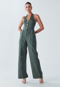 Zelený overal s halter výstřihem do V, se třemi zlatými knoflíky a širokými nohavicemi. Hladká textura a strukturovaný střih.