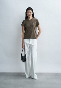 Καφέ κοντομάνικη μπλούζα με μικρό λογότυπο στο στήθος, σε συνδυασμό με λευκό ψηλόμεσο παντελόνι φαρδιάς γραμμής και μικρή μαύρη τσάντα.