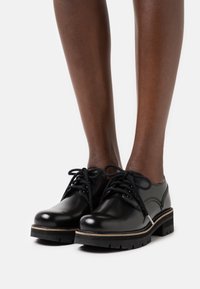 Chaussures en cuir noir à lacets avec une finition lisse, présentant une semelle épaisse et une bande contrastante plus claire le long du bord.