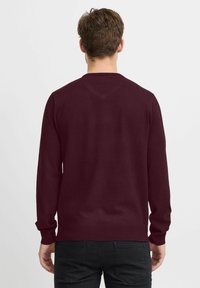 Mann iført en mørk rød langermet genser og svarte jeans, stående vendt bort fra kameraet mot en enkel hvit bakgrunn.