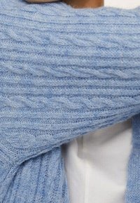 Pull tricoté bleu clair avec un motif torsadé, à la finition douce et texturée. Le vêtement est posé sur une chemise blanche à col.