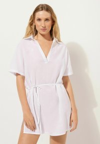 Vestido de lino blanco con cuello camisero, mangas cortas y cintura con cordón. Presenta un corte relajado y aberturas laterales para mayor libertad de movimiento.