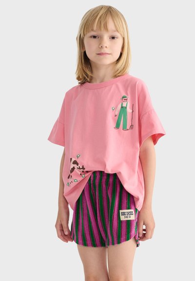 Enfant portant un t-shirt rose avec une illustration d'agriculteur et de vache, associé à un short rayé vert et violet avec un écusson de marque.