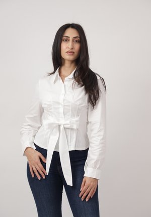 Femme debout aux longs cheveux bruns, portant une blouse blanche à boutons nouée à la taille et un jean bleu foncé, sur un fond uni.