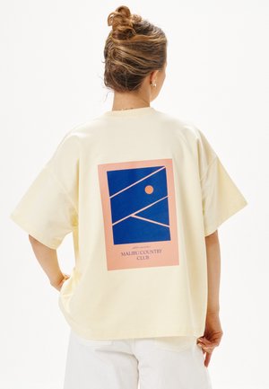 Frau mit Dutt, die ein lockeres cremefarbenes T-Shirt mit blauem und pinkem geometrischem Design und dem Text "Malibu Country Club" auf dem Rücken trägt.