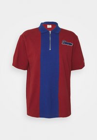 Polo rouge à manches courtes avec une bande verticale bleue centrale, col zippé et logo "Champion" sur la poitrine.