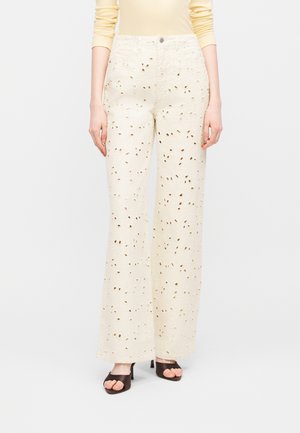 Γυναίκα που φοράει κρεμ παντελόνι με φαρδιά μπότα και λεπτομέρειες με κοψίματα, συνδυασμένο με μαύρες ανοιχτές γόβες και μακρυμάνικο μπεζ τοπ.