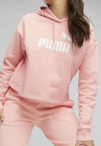 Roze cropped hoodie met een groot wit "PUMA" logo en bijpassende broek. Zacht materiaal, lange mouwen en een kangoeroezak.