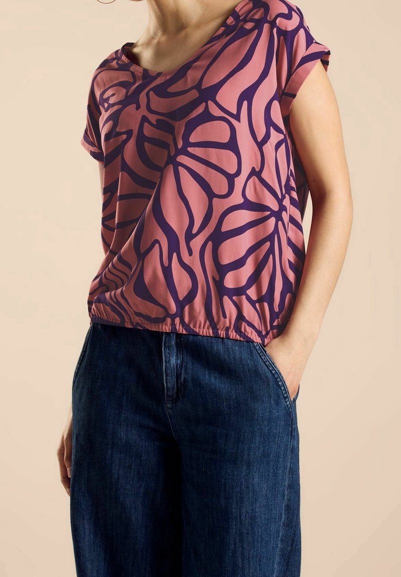 Short-sleeve top in pink with a dark purple floral pattern; flowy material and a gathered hem, paired with dark denim wide-leg pants.