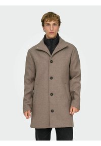 Cappotto marrone in misto lana con colletto a rever, chiusura frontale con bottoni e due tasche laterali. Dotato di uno strato interno nero rimovibile per maggiore calore.