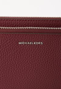 Cartera de cuero burdeos con acabado texturizado, que presenta un cierre de cremallera plateado y el logo "Michael Kors" visible en letras metálicas.
