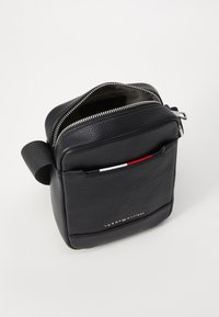 Schwarze Lederhandtasche mit strukturiertem Finish, Reißverschluss und Fronttasche mit roten und weißen Akzenten. Mit Markenlogo auf der Vorderseite.