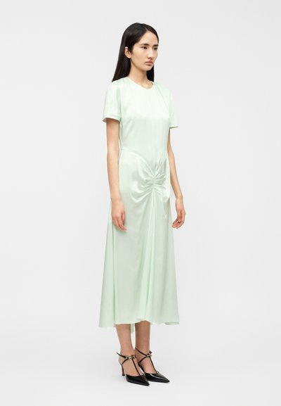 Φόρεμα από σατέν σε μέντα πράσινο με κοντά μανίκια, λεπτομέρεια σφικτού κόμπου στη μέση και φούστα μέχρι τους αστραγάλους. Φορεμένο με μαύρες γόβες μυτερές.