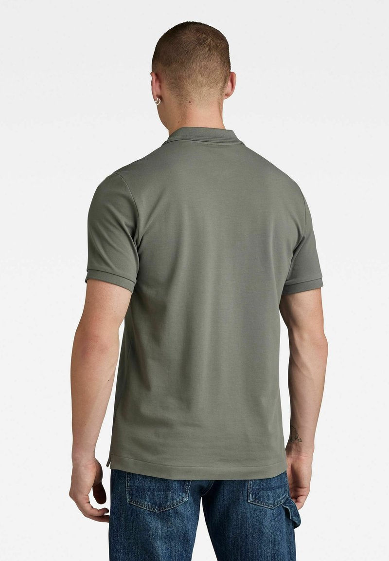 Olijfgroene polo met korte mouwen, gemaakt van gladde stof. Heeft een klassieke kraag en een rechte zoom, gecombineerd met donkerblauwe jeans.