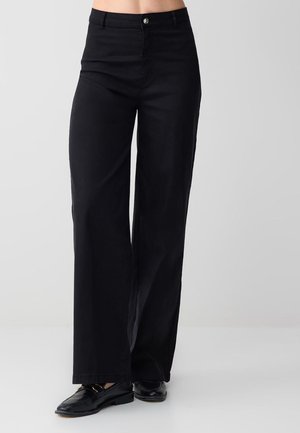 Femme portant un pantalon noir à taille haute et jambes larges ainsi que des mocassins noirs, debout devant un fond gris clair uni.