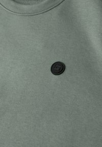 Sweat-shirt vert olive en tissu mélangé coton, avec un badge logo rond noir texturé sur la poitrine. Texture lisse, col rond.