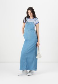 Abito maxi in denim con bretelle, colore azzurro chiaro, taglio dritto, tasche frontali, abbinato a una maglietta a righe e sneakers bianche.