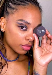 Zwart crèmekleurig oogschaduwcontainer met het etiket "963" dicht bij het oog gehouden. Model met een ingevlochten haarstijl, een kralenketting en lichte make-up.