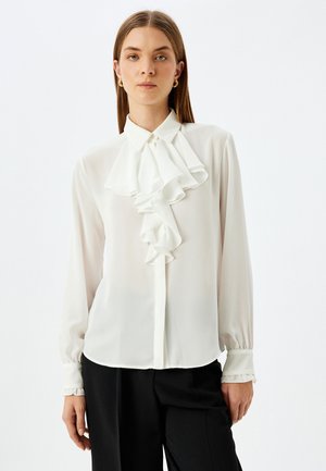 Blusa blanca con frente y cuello con volantes, mangas largas con puños fruncidos, hecha de una tela ligera, combinada con pantalones negros ajustados.