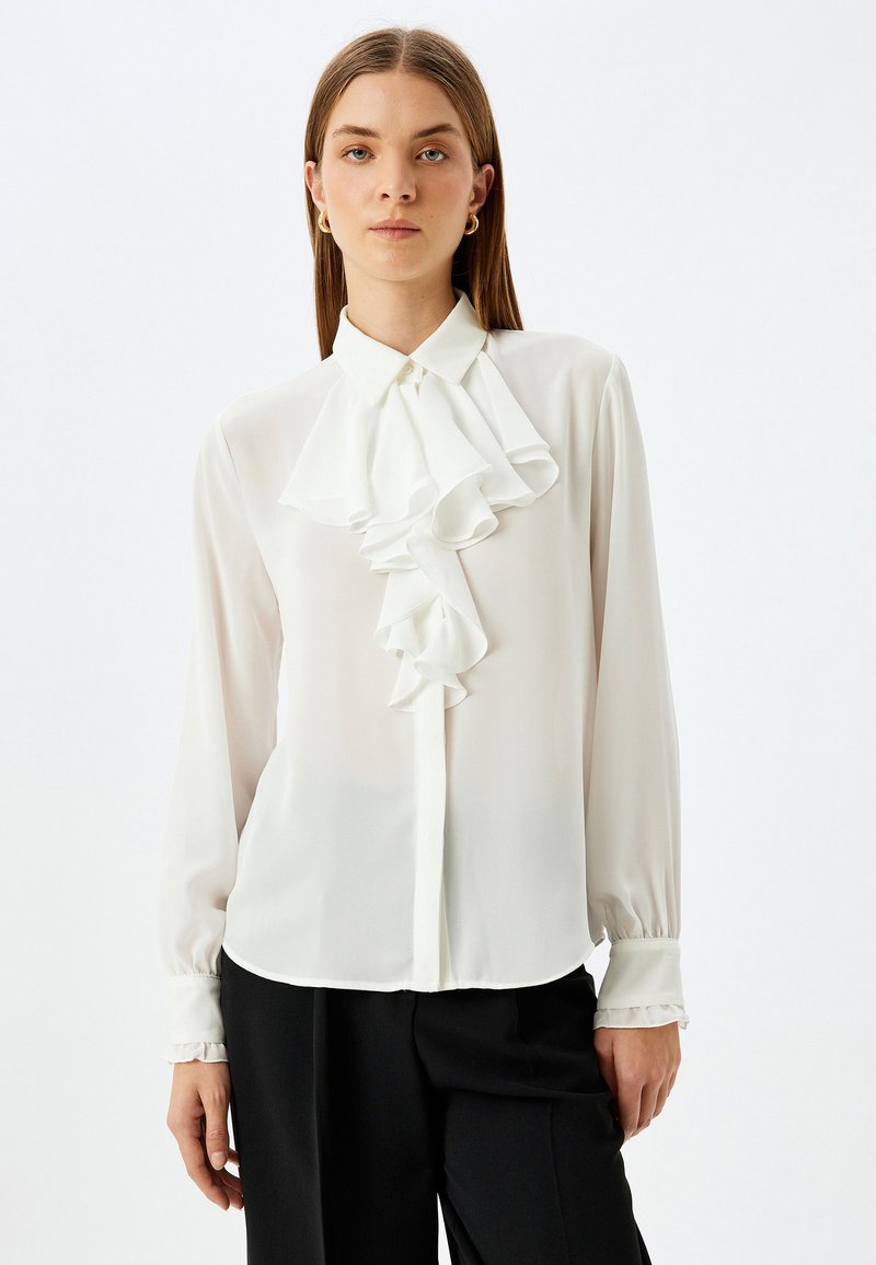 Blusa blanca con frente y cuello con volantes, mangas largas con puños fruncidos, hecha de una tela ligera, combinada con pantalones negros ajustados.